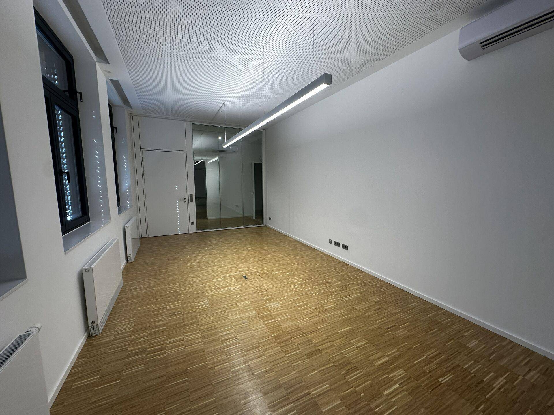 Moderner Büroraum mit Holzparkett, weißen Wänden, Heizkörpern und einer eleganten Glasabtrennung.