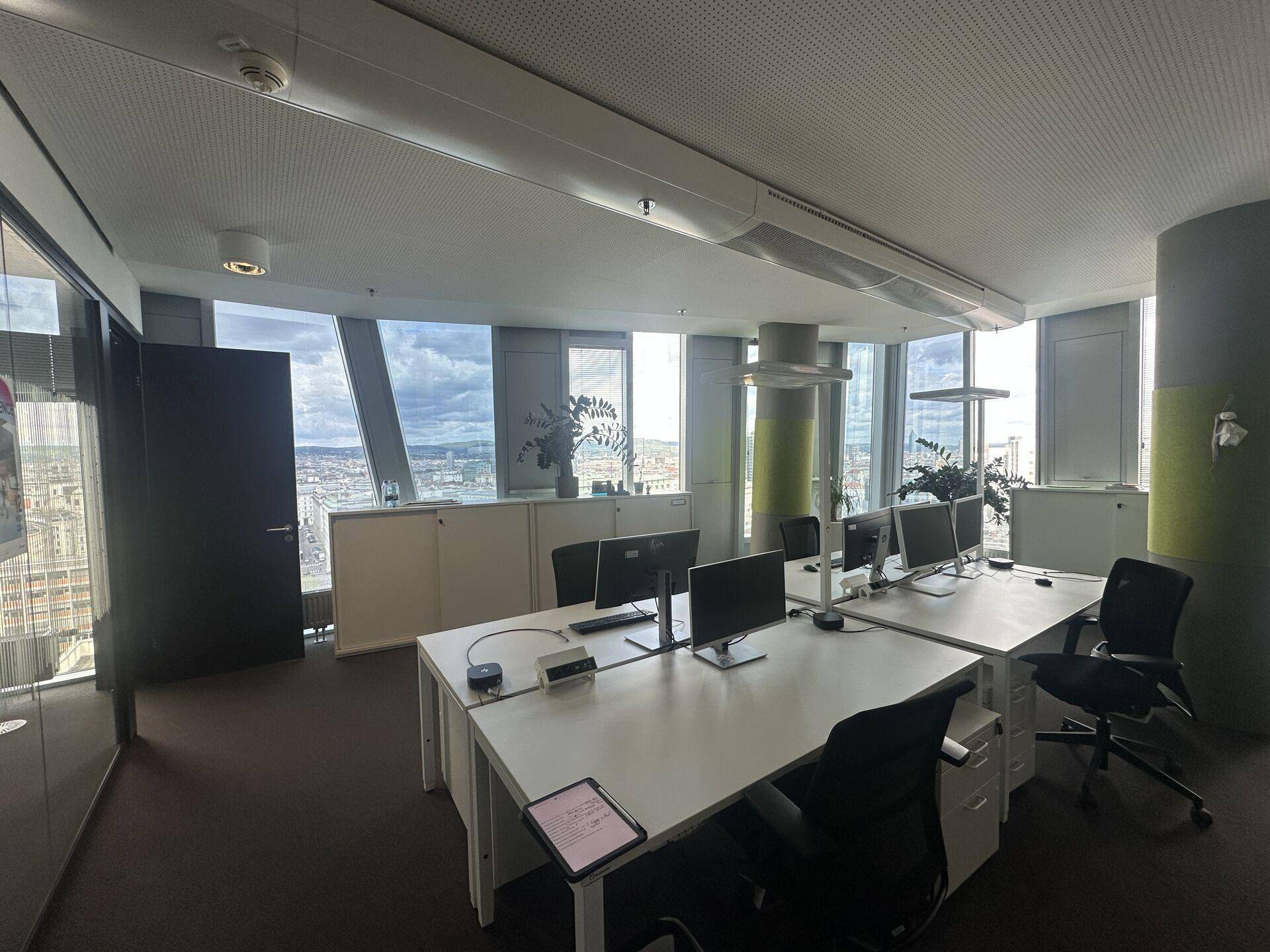 Modernes Großraumbüro mit mehreren Arbeitsplätzen, Computern und weitem Blick über die Stadt durch große Fenster.