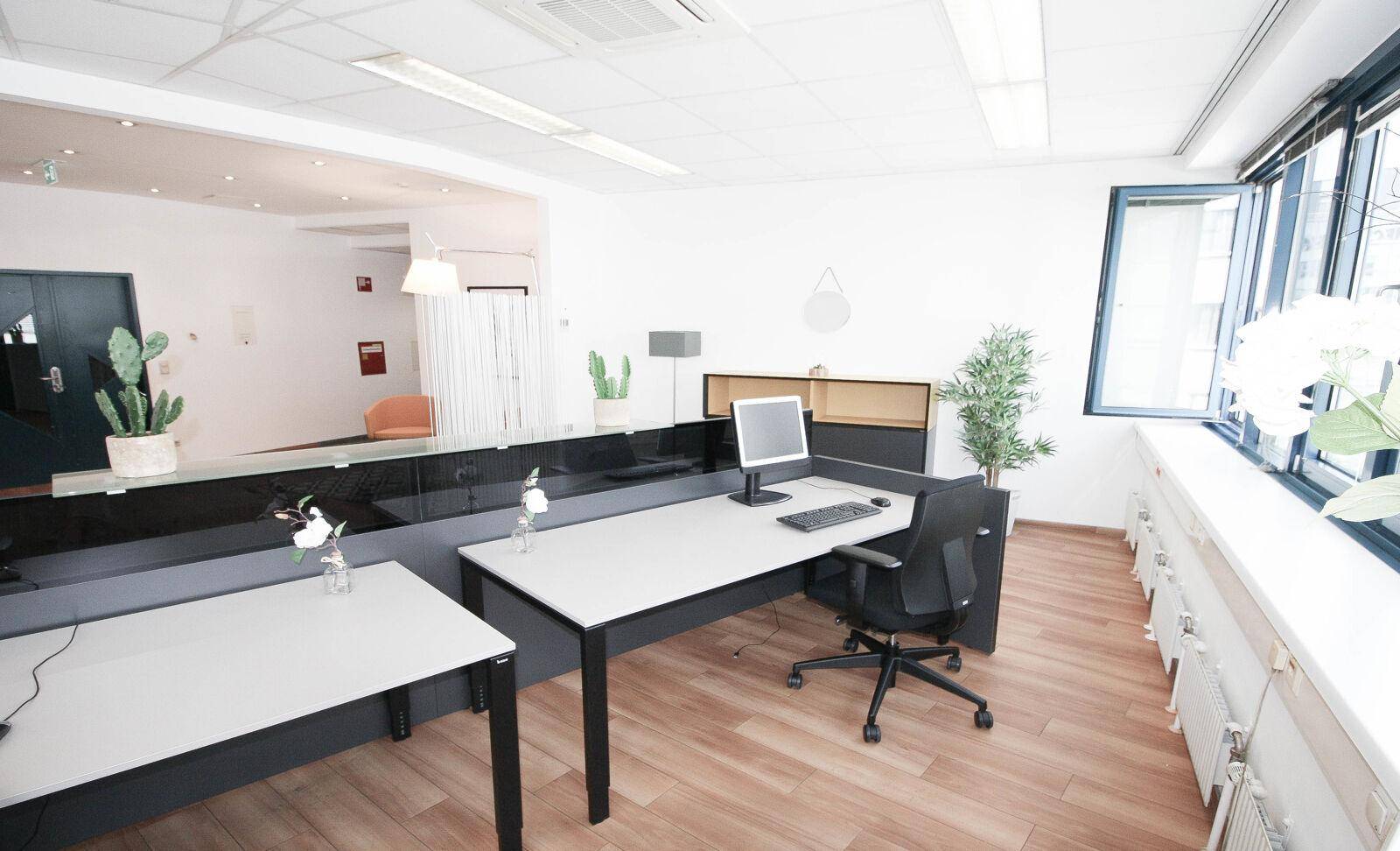 Offener Bürobereich mit Holzfußboden, modernen Schreibtischen und großen Fenstern, die viel Tageslicht hereinlassen.