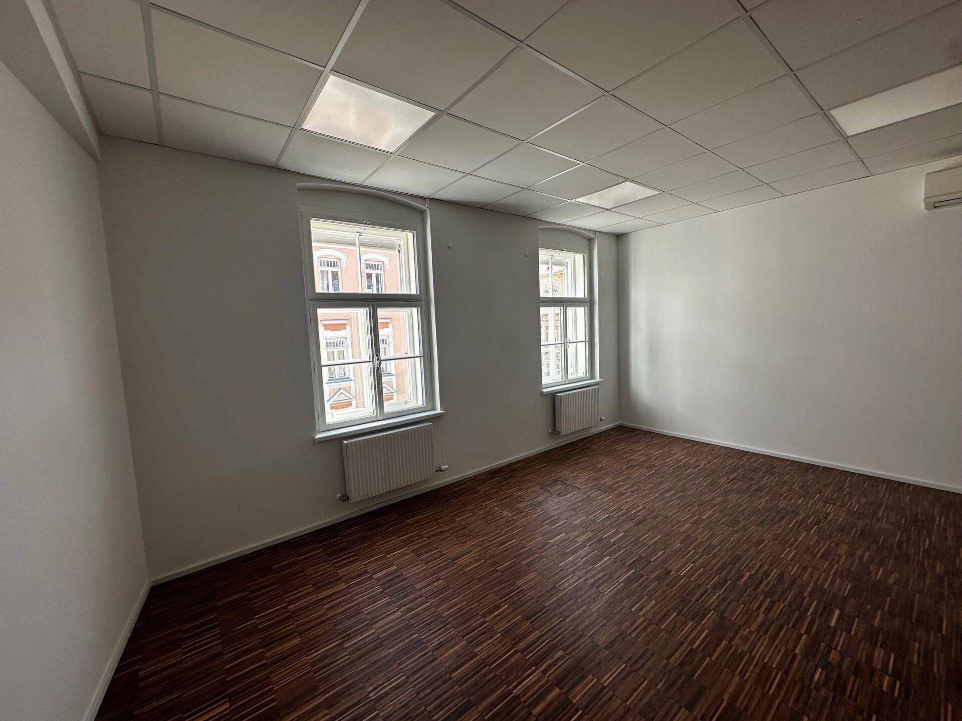 Geräumiges, leeres Büro mit dunklem Holzboden, weißen Wänden und zwei großen Fenstern.