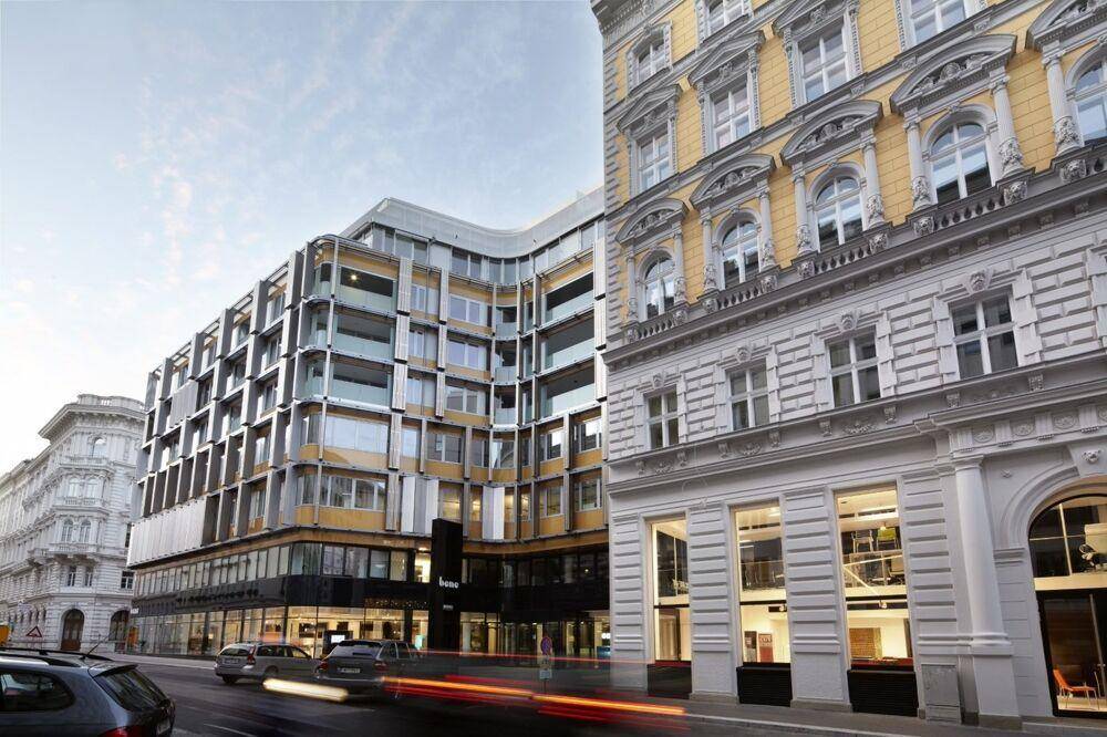 Moderne und historische Gebäudefronten entlang einer belebten Stadtstraße mit vorbeifahrenden Autos.