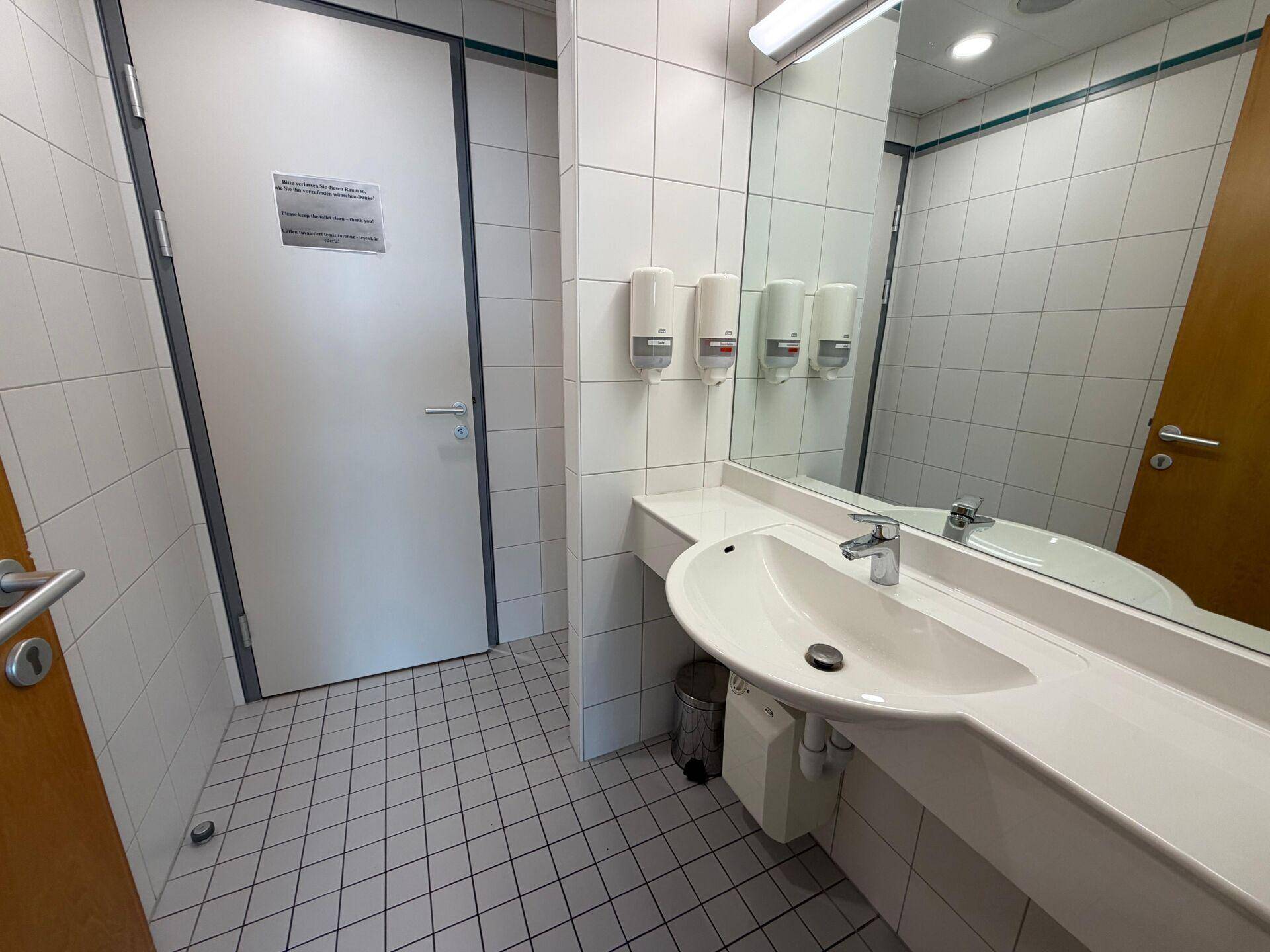 Einfaches Badezimmer mit Fliesenboden, Waschbecken, Spiegel und weißer Tür.