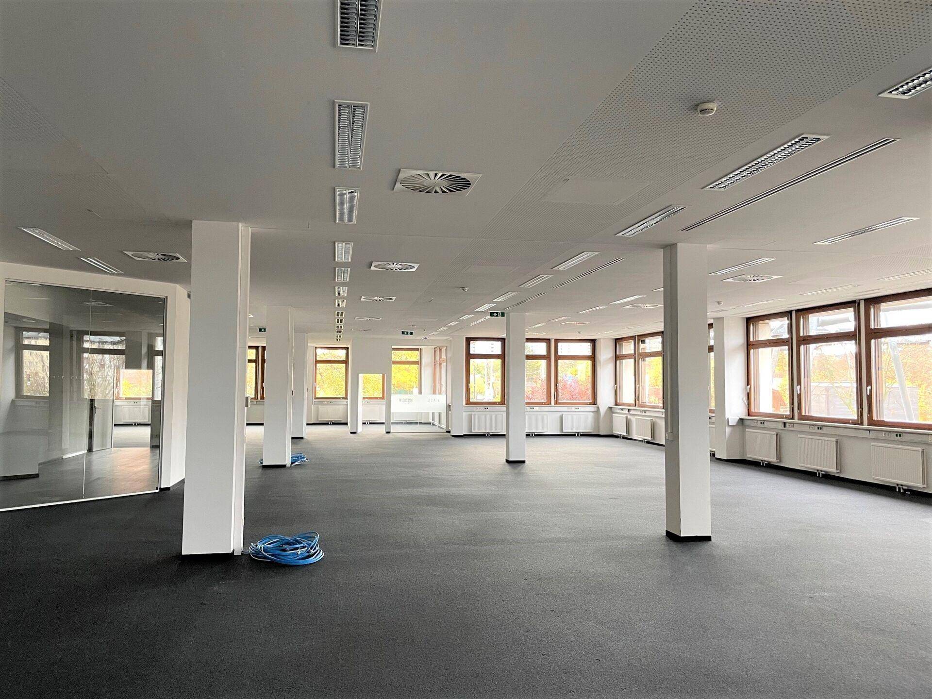 Großer, offener Büroraum mit dunklem Boden, weißen Säulen und vielen Fenstern, die viel Tageslicht hereinlassen.