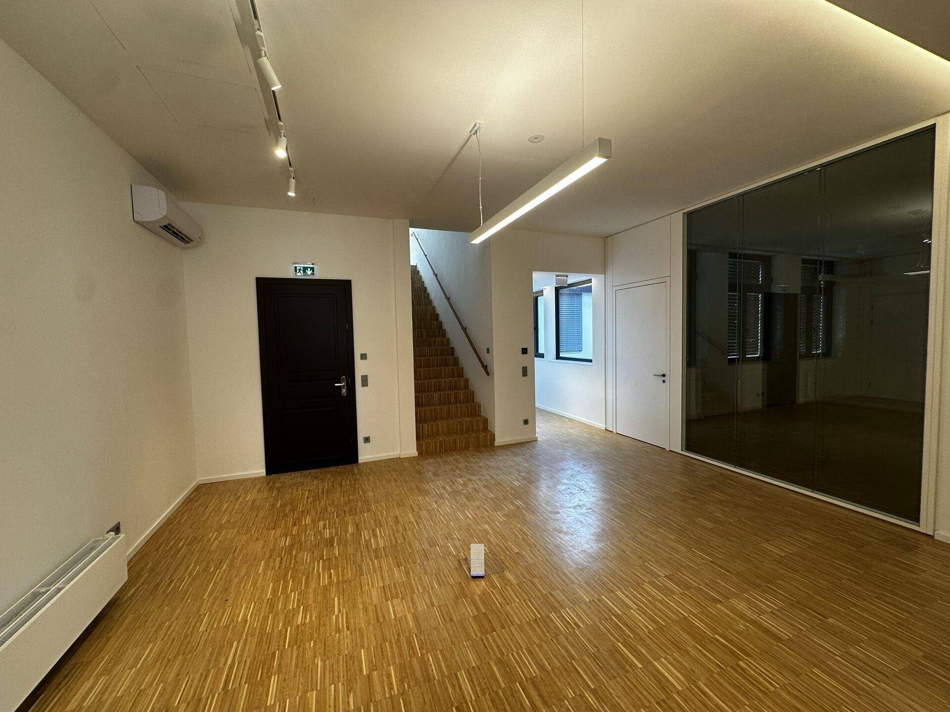 Geräumiger Bürobereich mit Holzparkett, moderner Beleuchtung, Treppenzugang und einer eleganten Glaswand.