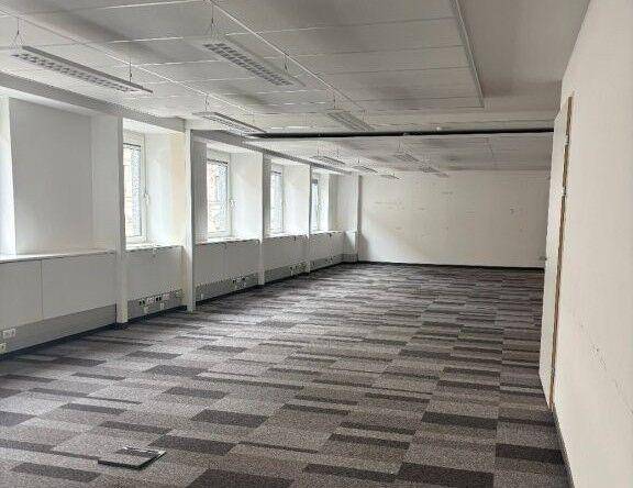 Geräumiges, leeres Büro mit Teppichboden und einer Reihe von Fenstern, die viel Tageslicht hereinlassen.