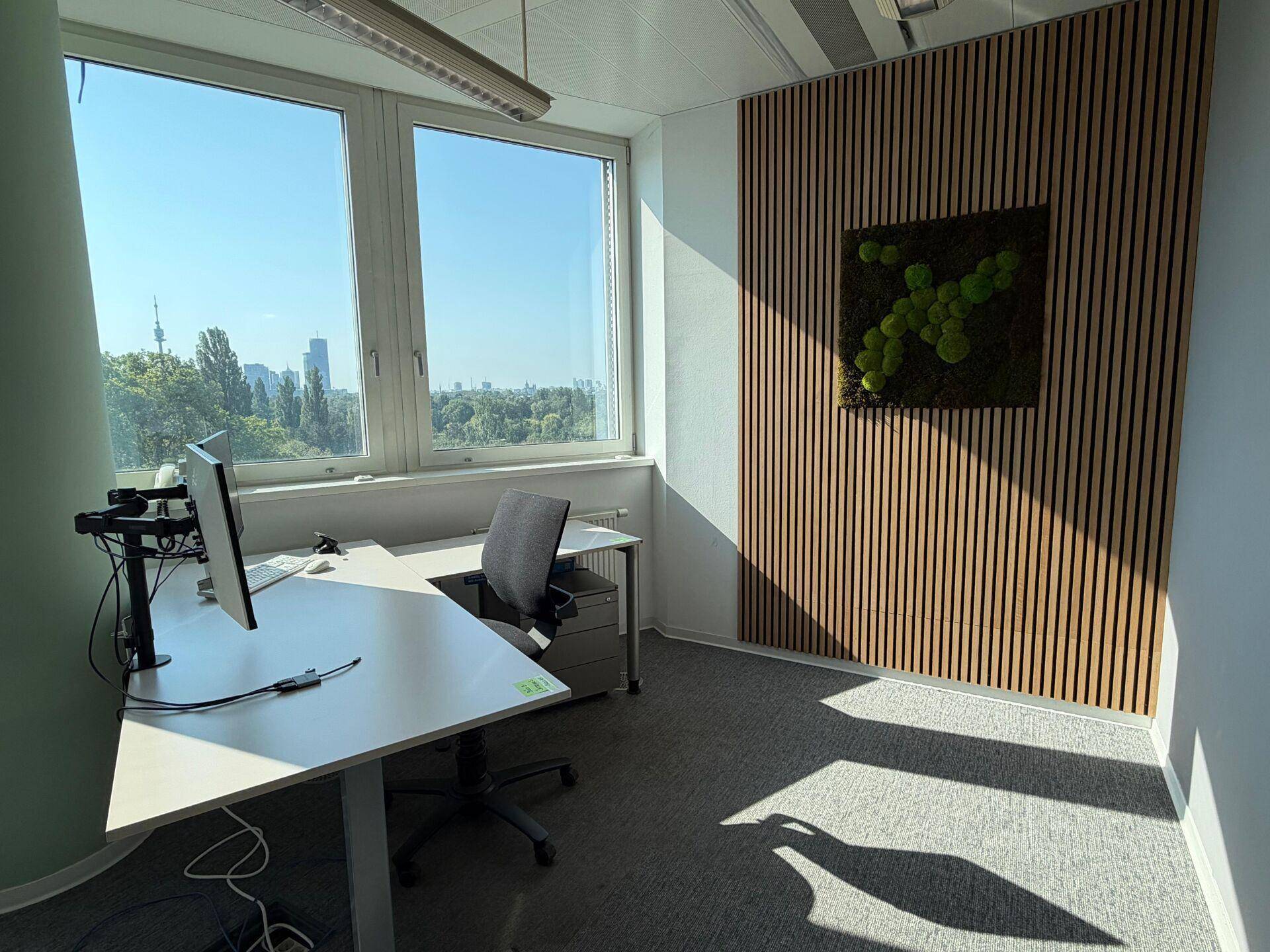 Helles Einzelbüro mit Schreibtisch, großen Fenstern und einer stilvollen Holzwand mit grüner Moosdekoration.