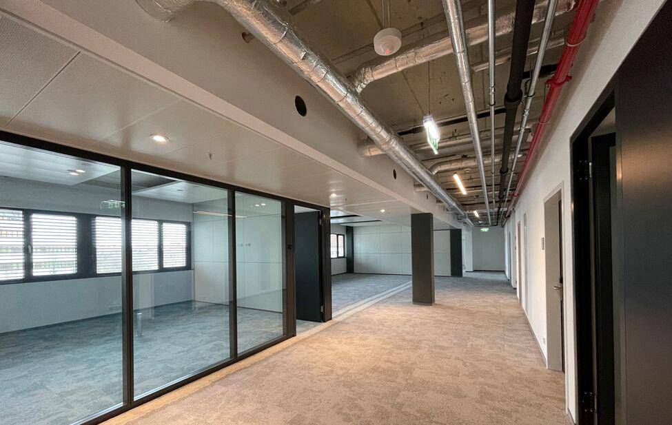 Geräumiger Bürobereich mit Teppichboden, Glaswänden und sichtbaren Lüftungsrohren an der Decke.