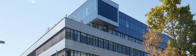Moderne Bürogebäude-Fassade mit großen Glasflächen, blauer Himmelsreflexion und grünen Bäumen.