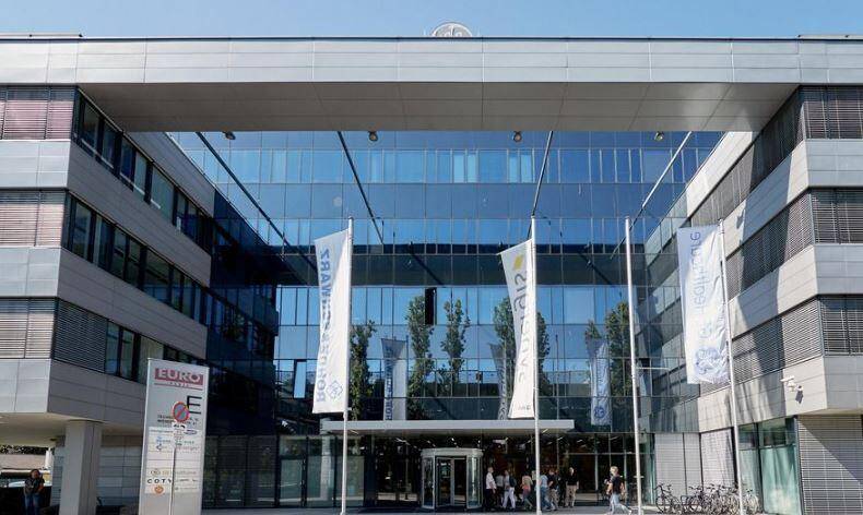Eingangsbereich eines modernen Bürogebäudes mit beeindruckender Glasfassade, Drehtür und Firmenflaggen.