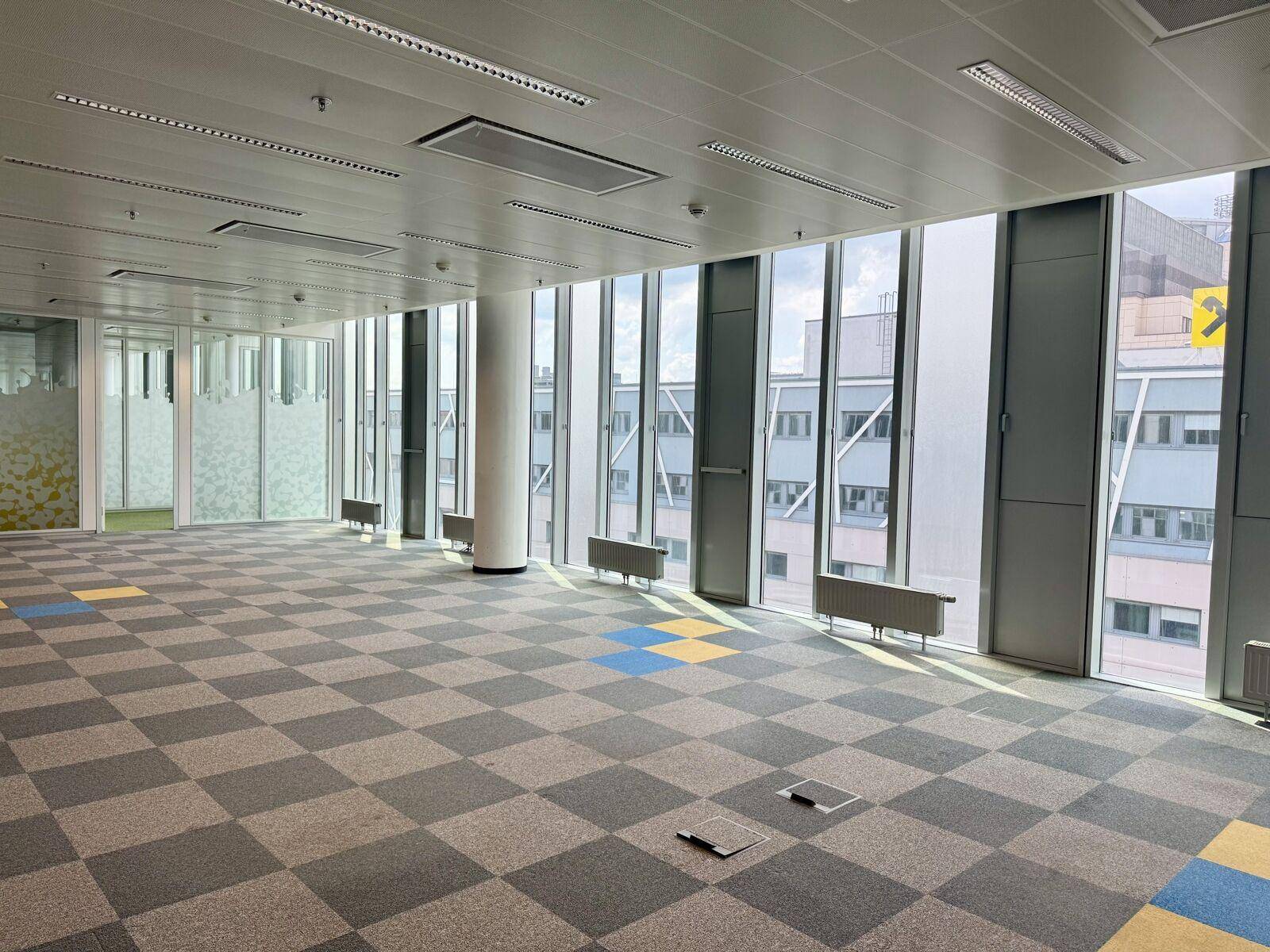 Großes, leeres Büro mit kariertem Teppichboden, raumhohen Fenstern und Heizkörpern unter den Fenstern.