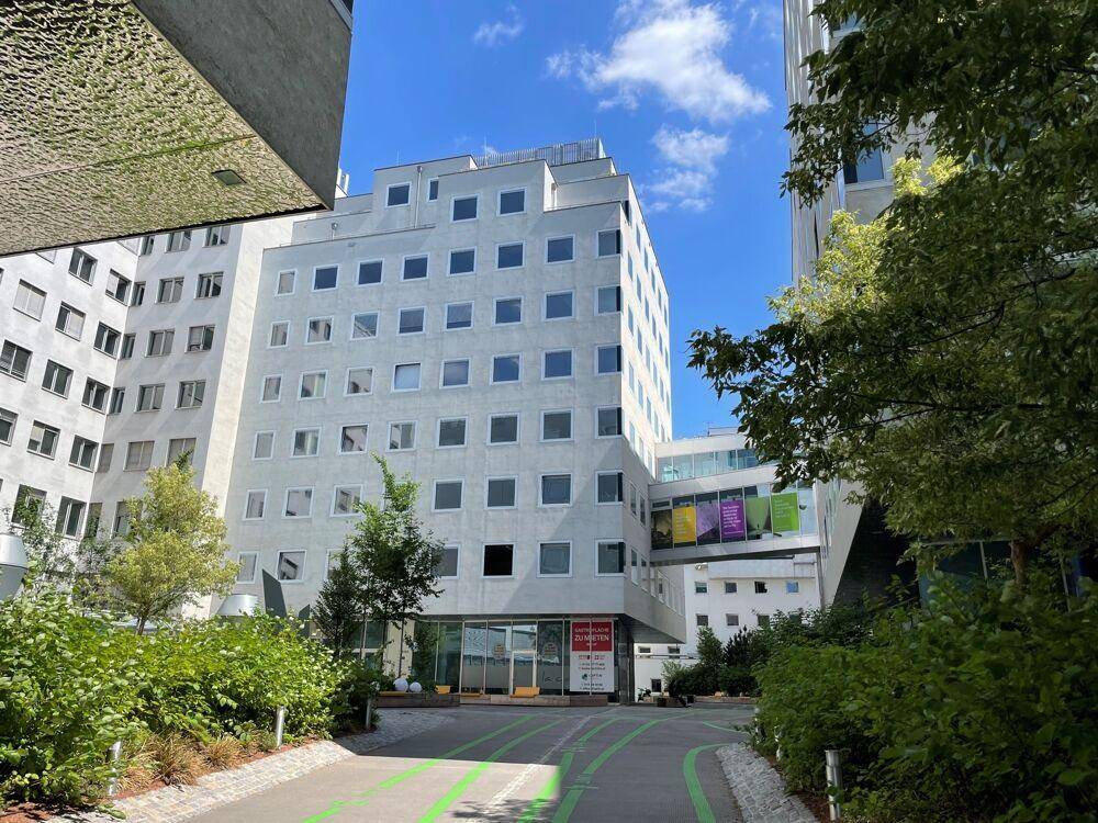 Modernes Gebäudeensemble mit weißer Fassade, vielen Fenstern und begrünten Außenbereichen unter blauem Himmel.