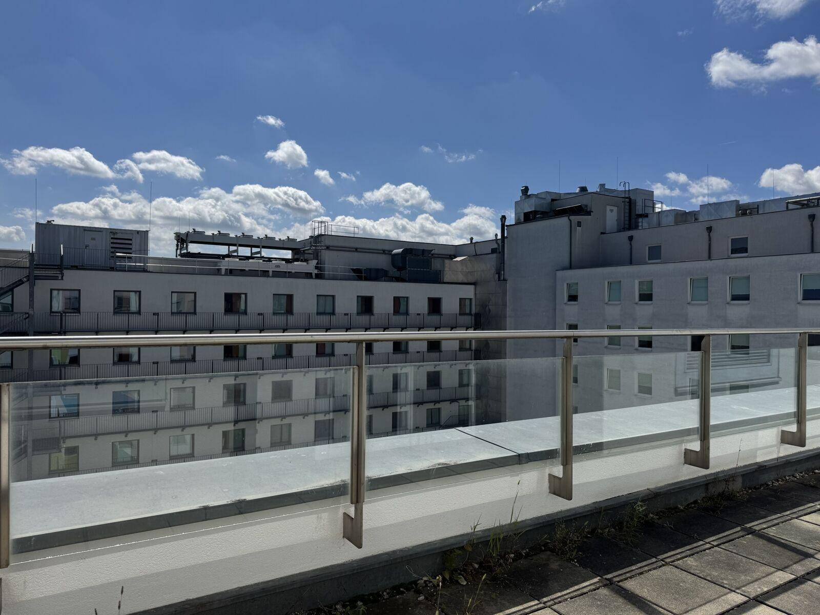 Terrasse mit Glasgeländer und Blick auf umliegende Gebäude unter blauem Himmel mit Wolken.
