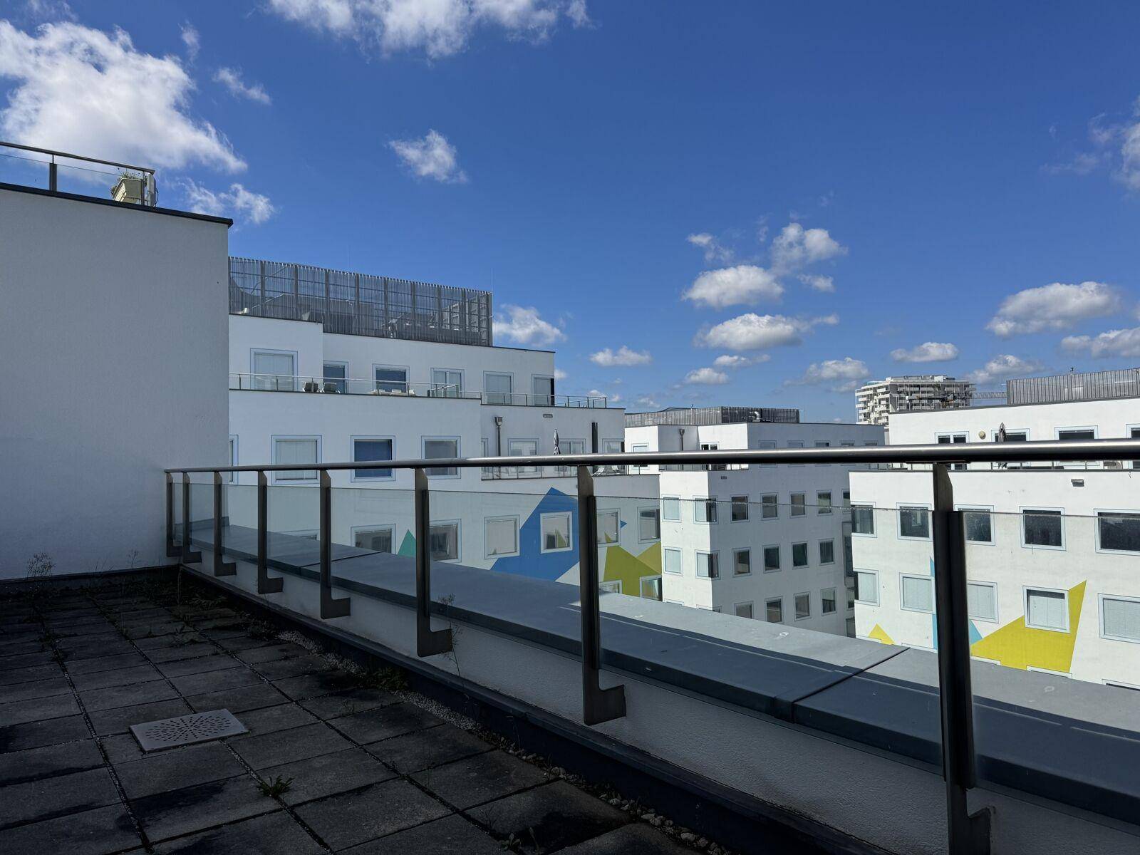 Terrasse mit Glasgeländer und Blick auf umliegende moderne Gebäude mit farbigen Akzenten unter blauem Himmel.