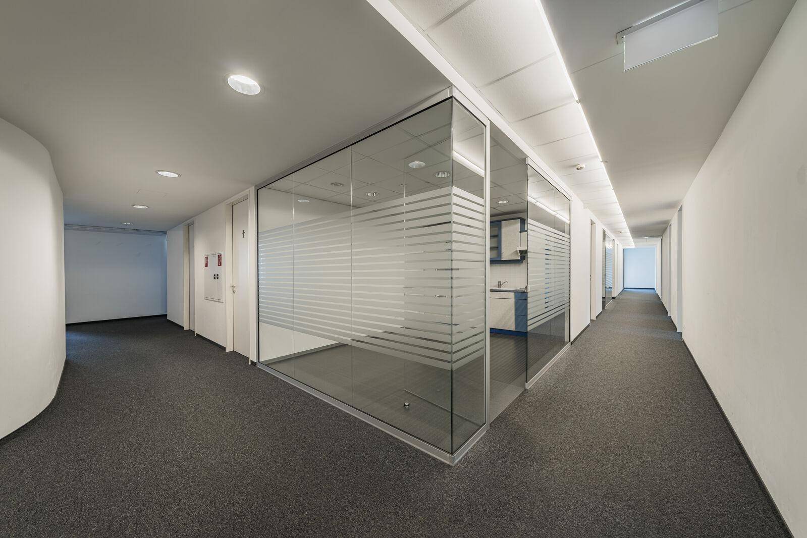 Moderner Büroflur mit Glaswänden zu den Büros und einer kleinen Teeküche im Hintergrund.