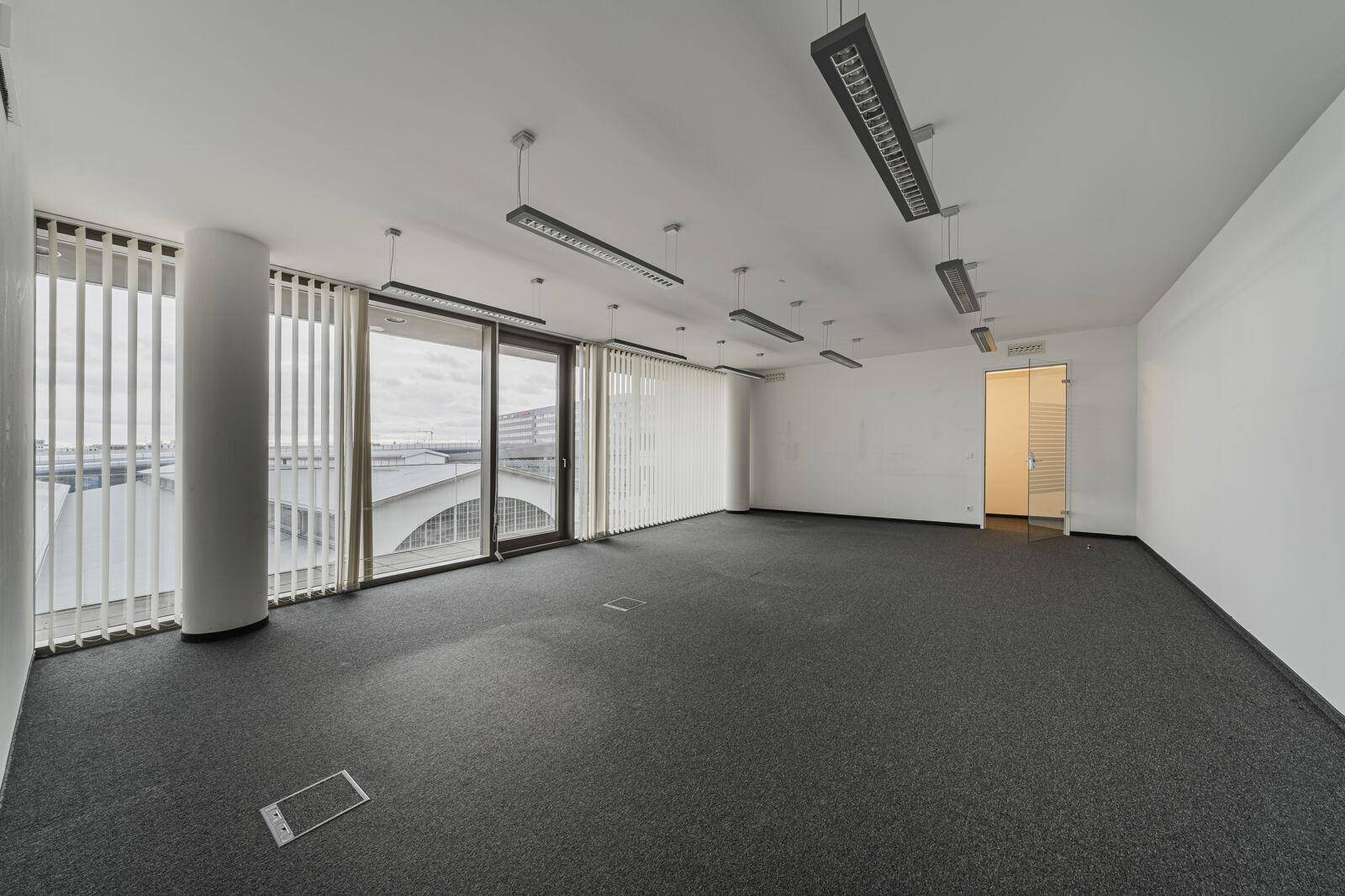 Geräumiger, leerer Büroraum mit grauem Teppichboden, großen Fenstern mit Jalousien und einer Glastür.