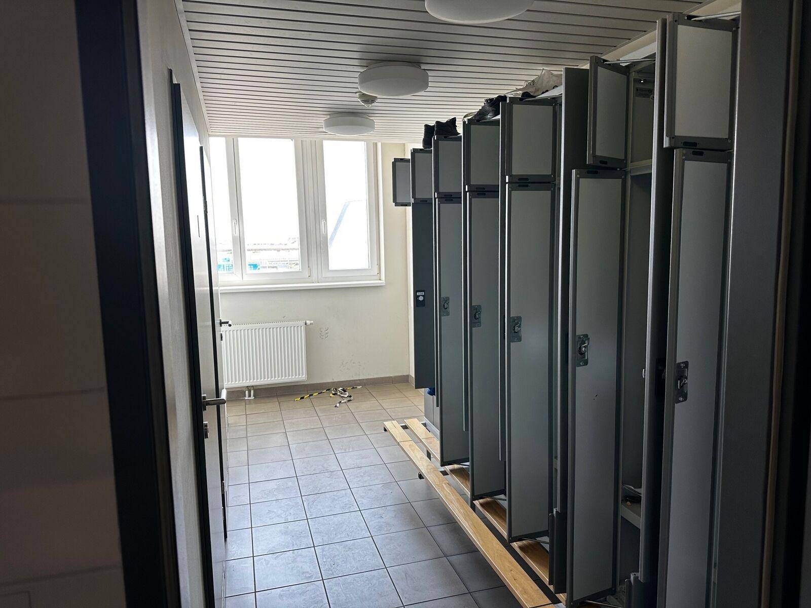 Umkleideraum mit Reihen von Spinden, Fliesenboden und einem Fenster, gut beleuchtet.