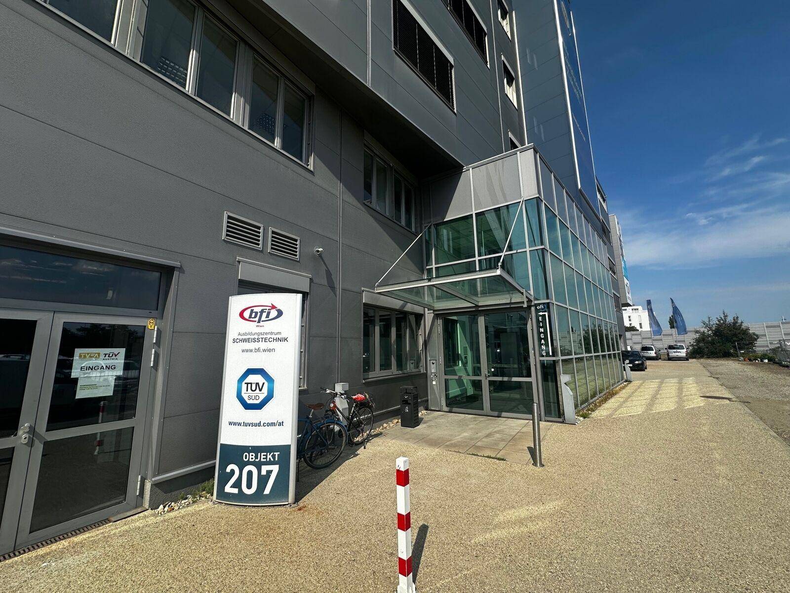 Eingangsbereich eines modernen Bürogebäudes mit Glasfassade und Beschilderung unter blauem Himmel.