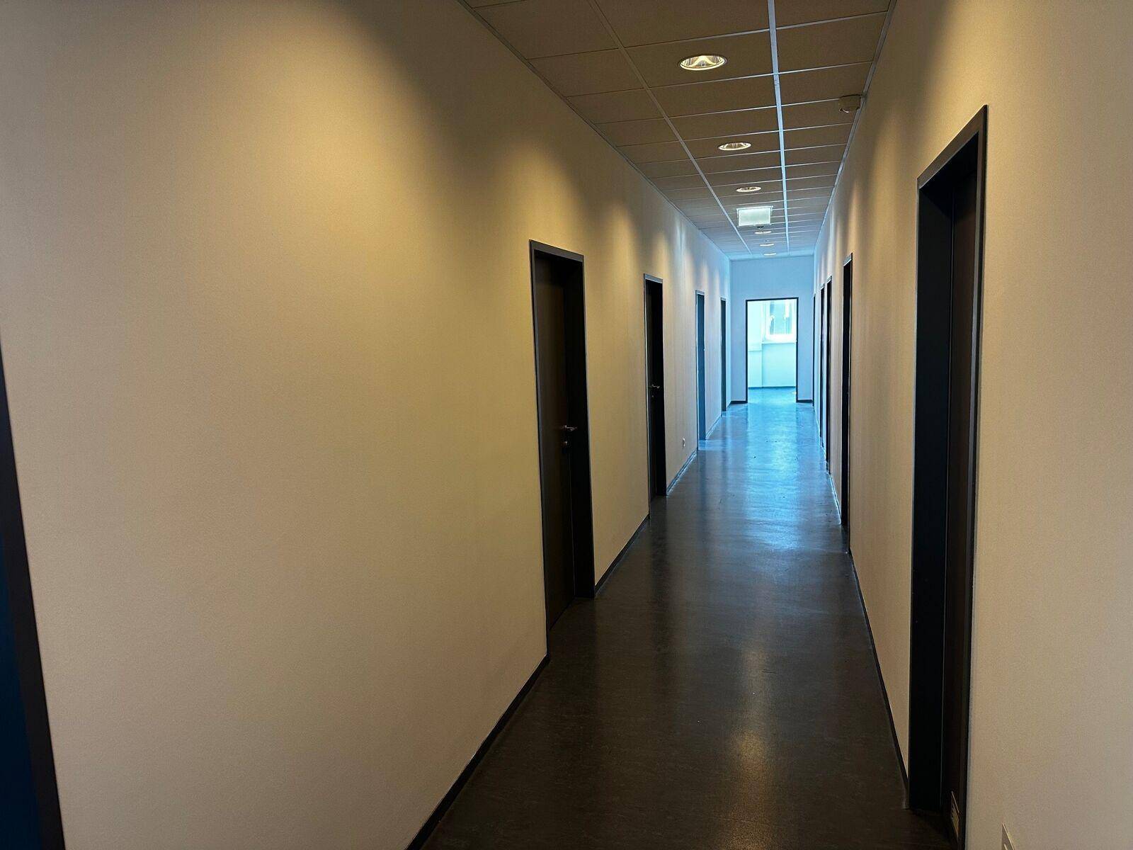 Langer Flur mit mehreren Türen, abgehängter Decke und dunklem Bodenbelag, gut beleuchtet.