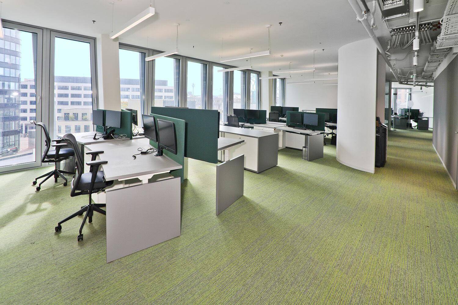 Moderner Großraumbüro mit ergonomischen Arbeitsplätzen, grünen Teppichböden und großen Fenstern.