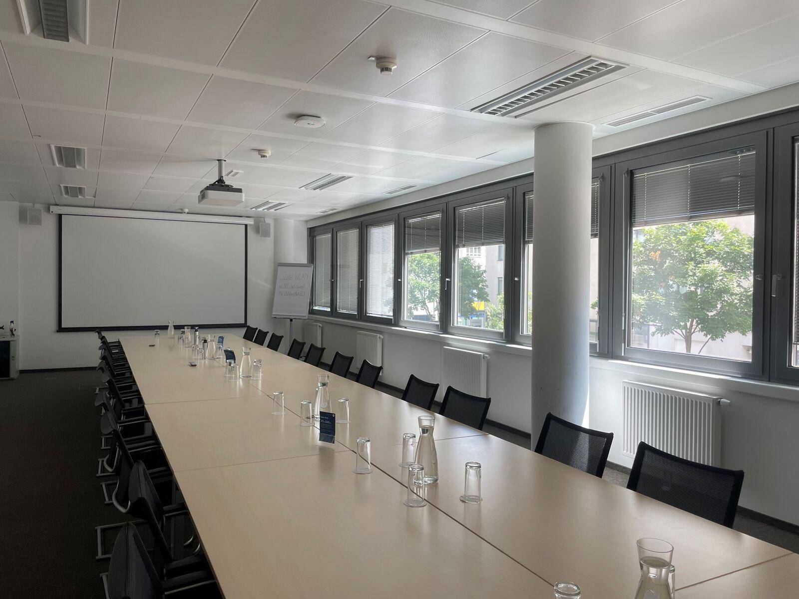 Großer Konferenzraum mit langem Tisch, vielen Stühlen, Beamer und großen Fenstern.