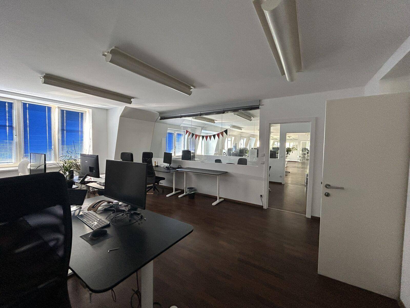 Großes, helles Büro mit mehreren Arbeitsplätzen, dunklem Laminatboden und blauen Jalousien an den Fenstern.