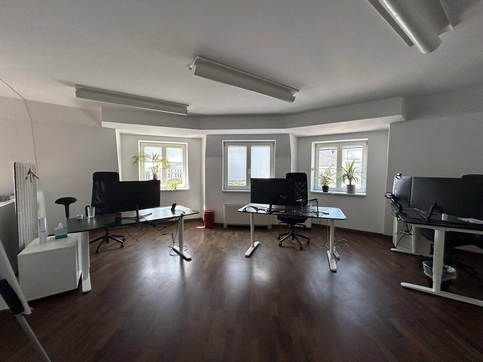 Helles Eckbüro mit mehreren Fenstern, dunklem Laminatboden und modernen Arbeitsplätzen mit Blick ins Grüne.