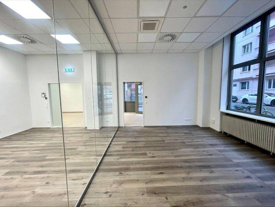 Heller Gewerberaum mit Holzoptik-Boden, großen Fenstern und einer prominenten Spiegelwand.