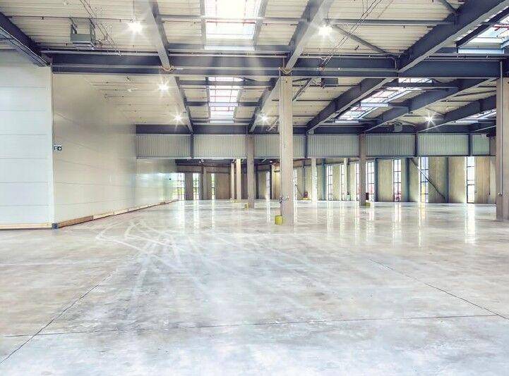 Helle, leere Lagerhalle mit poliertem Betonboden, hohen Decken und natürlichem Lichteinfall durch Oberlichter.