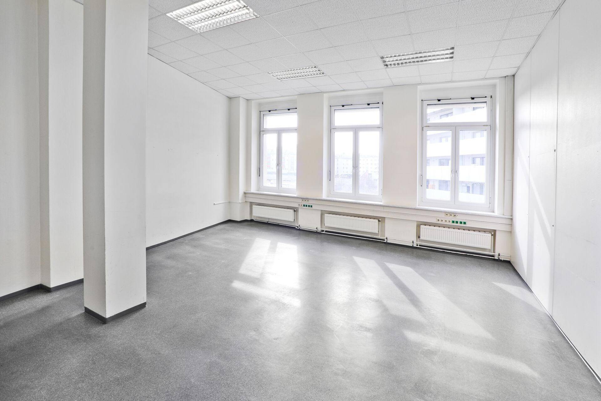 Leerer Büroraum mit großen Fenstern, Säulen und einem modernen grauen Bodenbelag.
