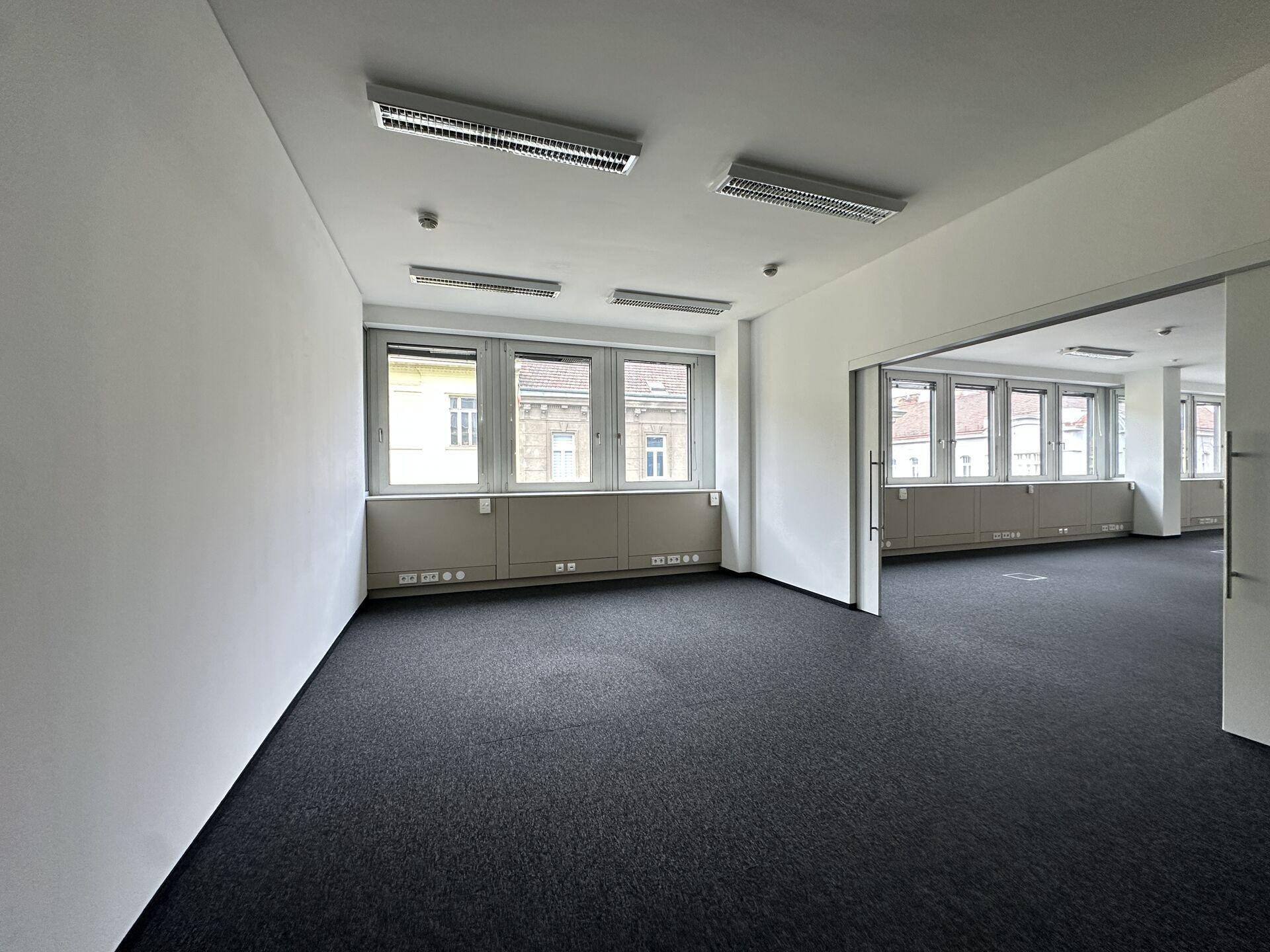 Leerer Büroraum mit dunklem Teppichboden, weißen Wänden und mehreren Fenstern mit Blick nach draußen.