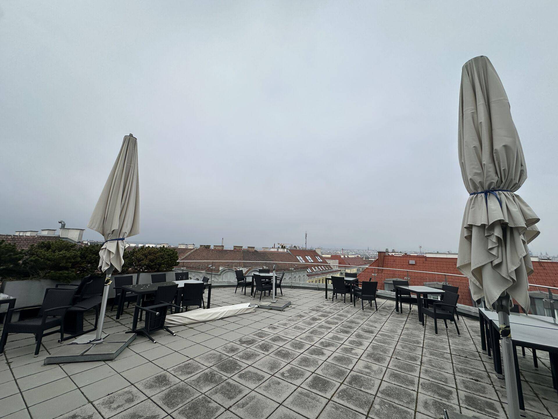 Große Dachterrasse mit Tischen, Stühlen und zusammengeklappten Sonnenschirmen unter bewölktem Himmel.