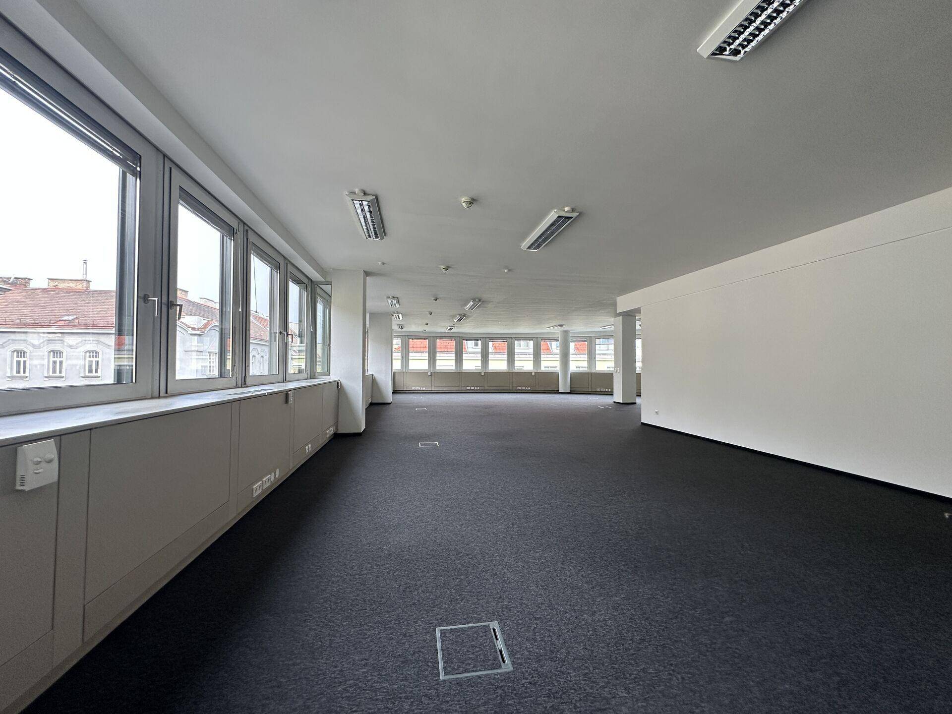 Geräumiger, leerer Bürobereich mit durchgehenden Fenstern, dunklem Teppich und weißen Wänden.