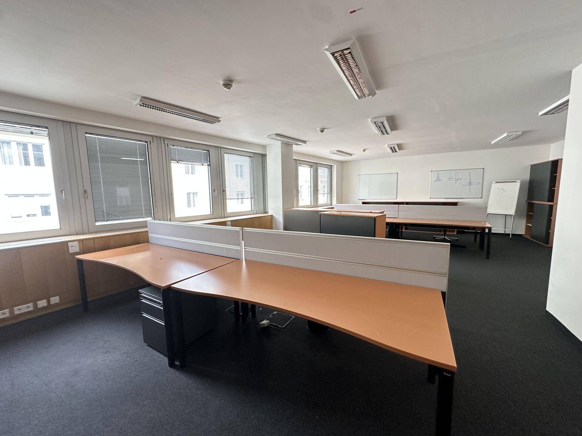 Büroraum mit mehreren Schreibtischen, Trennwänden, dunklem Teppichboden und großen Fenstern.