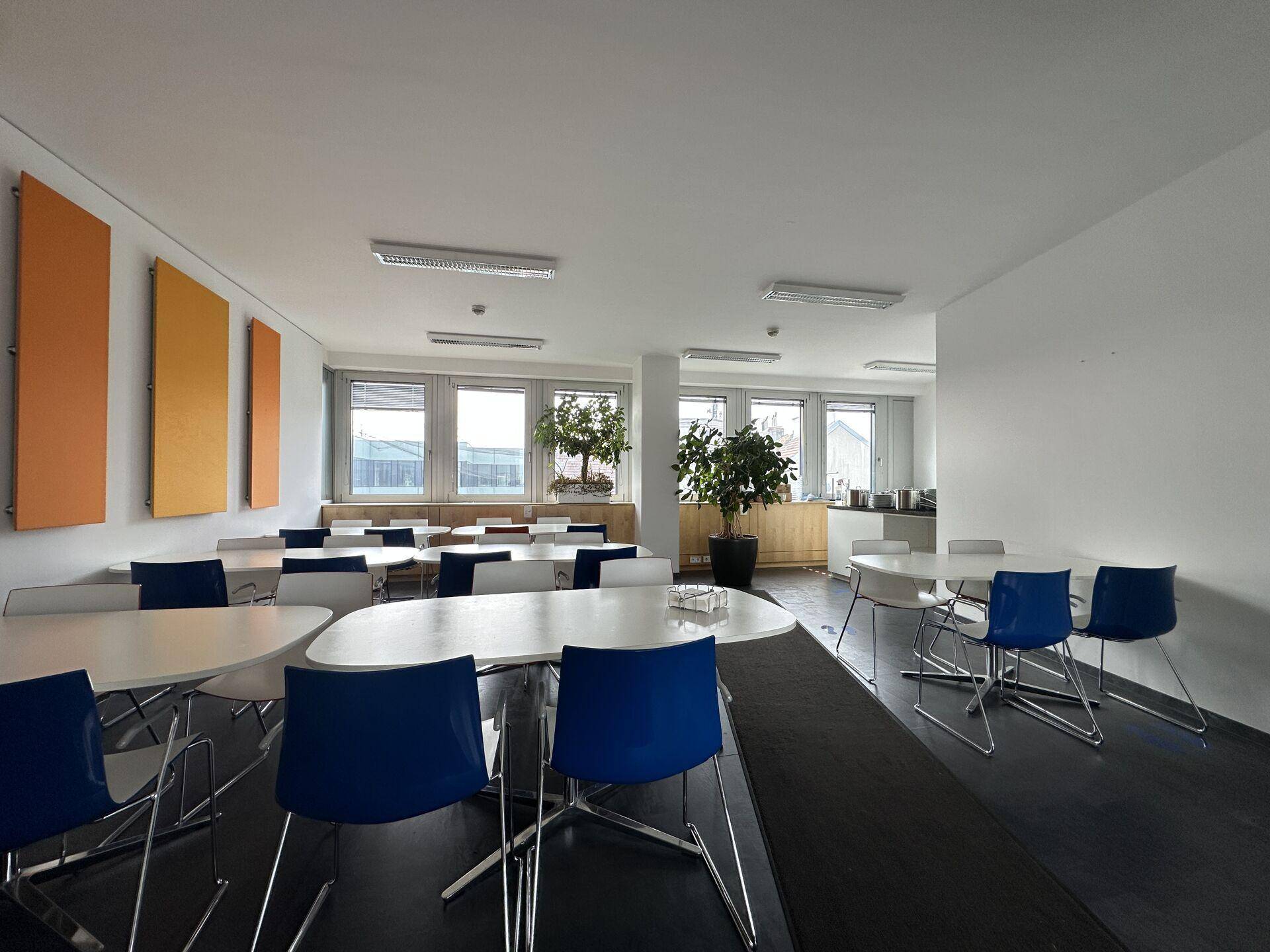 Helle Kantine mit vielen Tischen und blauen Stühlen, großen Fenstern und orangefarbenen Akzenten.