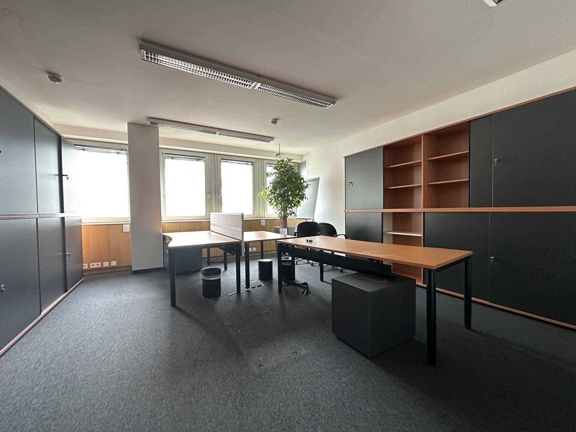 Büro mit Schreibtischen, Schränken, Teppichboden und Fenstern mit Tageslicht.