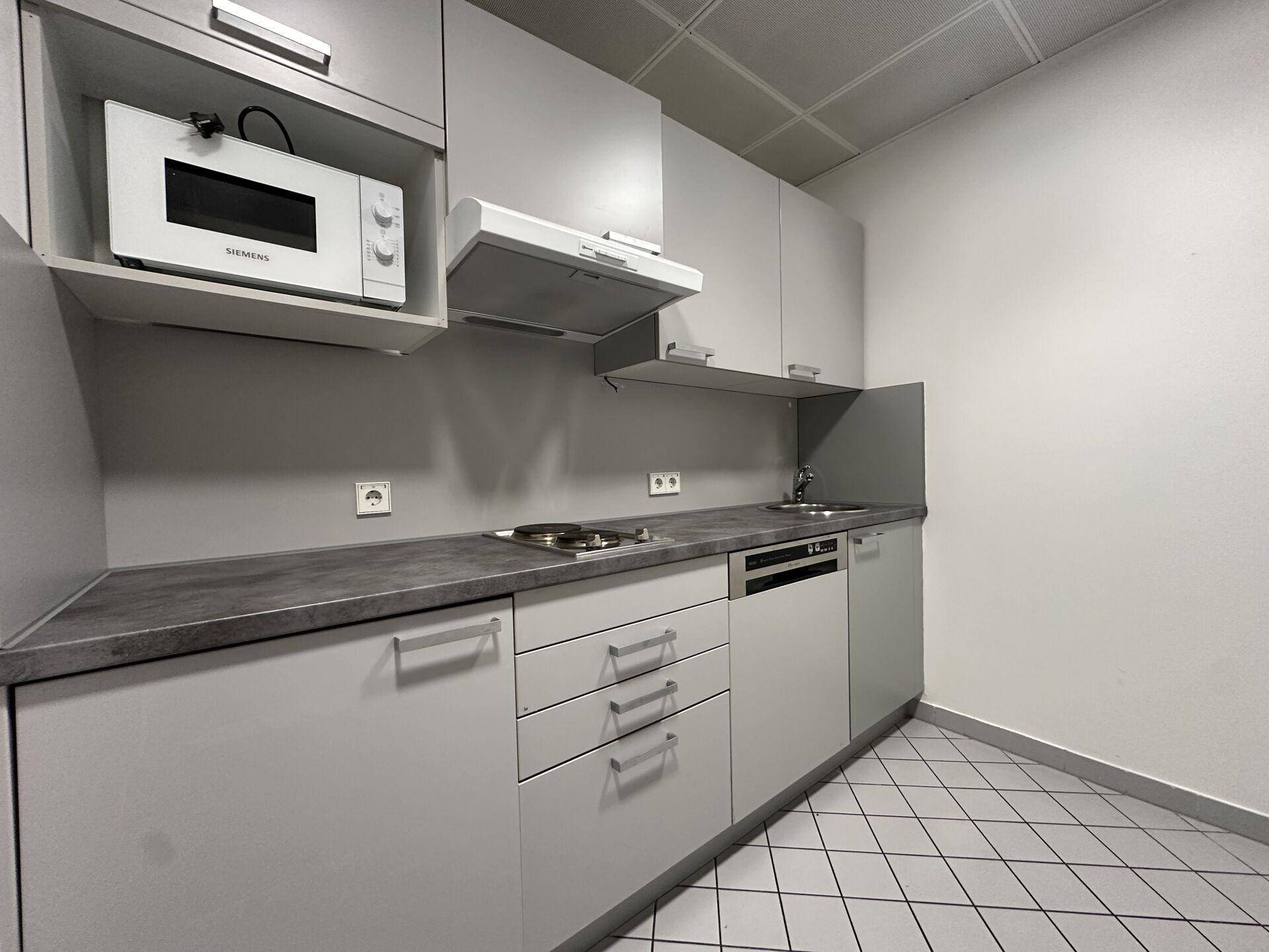 Kleine Teeküche mit grauen Schränken, Mikrowelle, Kochfeld und Spülmaschine, gefliestem Boden.