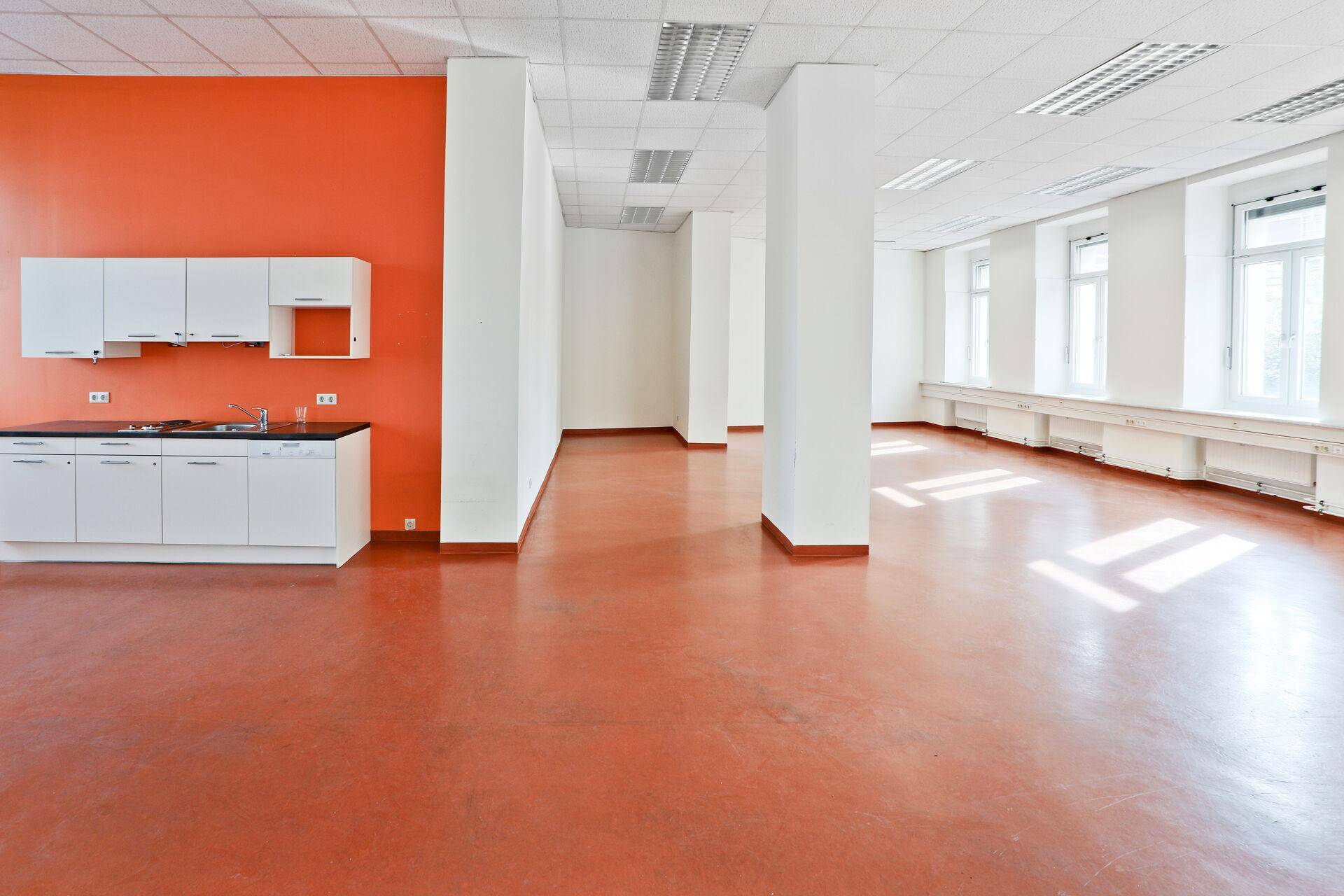 Geräumiger Büroraum mit orangefarbener Akzentwand, kleiner Küchenzeile und glänzendem Boden.
