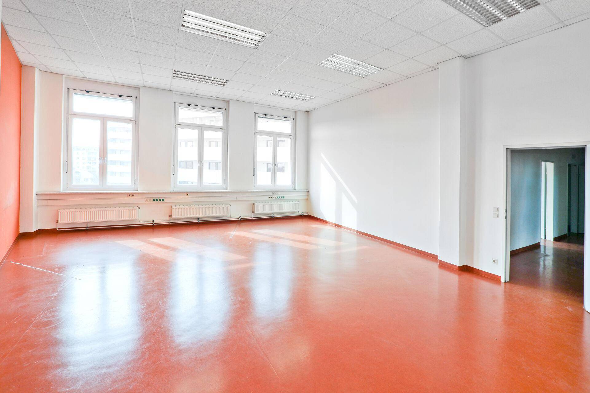 Heller Büroraum mit großen Fenstern, viel Tageslicht und einem glänzenden, rötlich-braunen Boden.