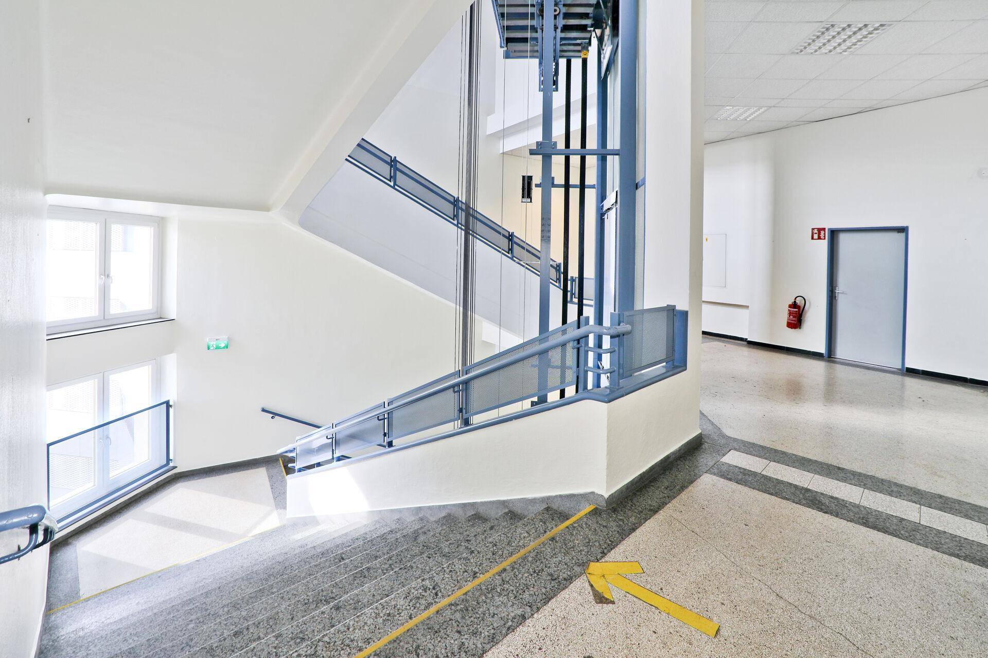 Heller Treppenaufgang mit modernem Glasaufzug und Terrazzoboden in einem Bürogebäude.