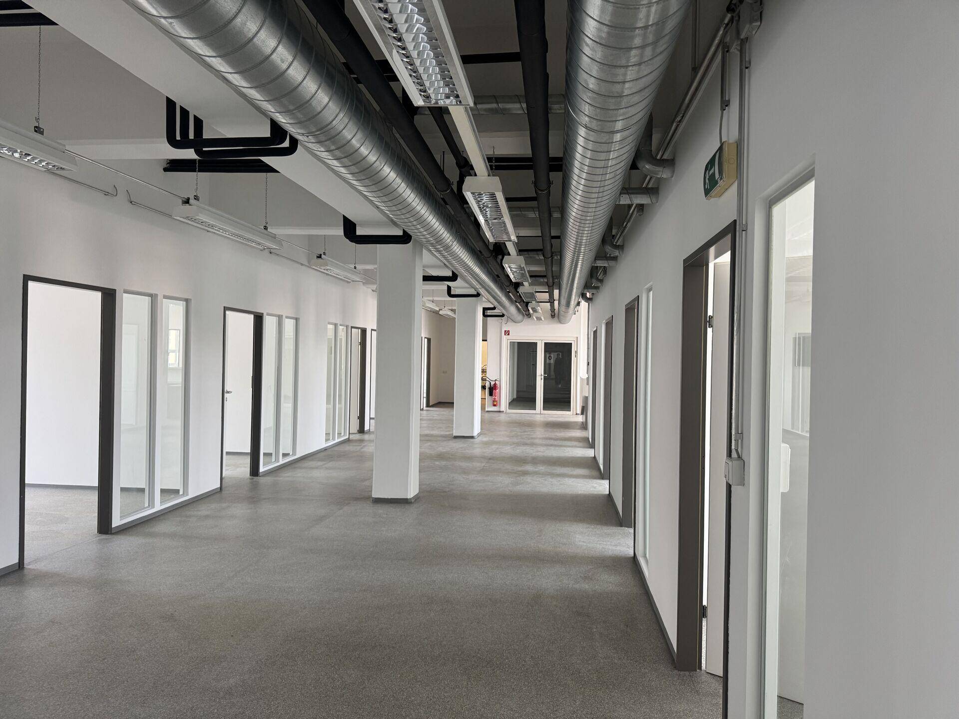 Langer, leerer Büroflur mit sichtbaren Lüftungsrohren und vielen Bürotüren mit Glasfronten.