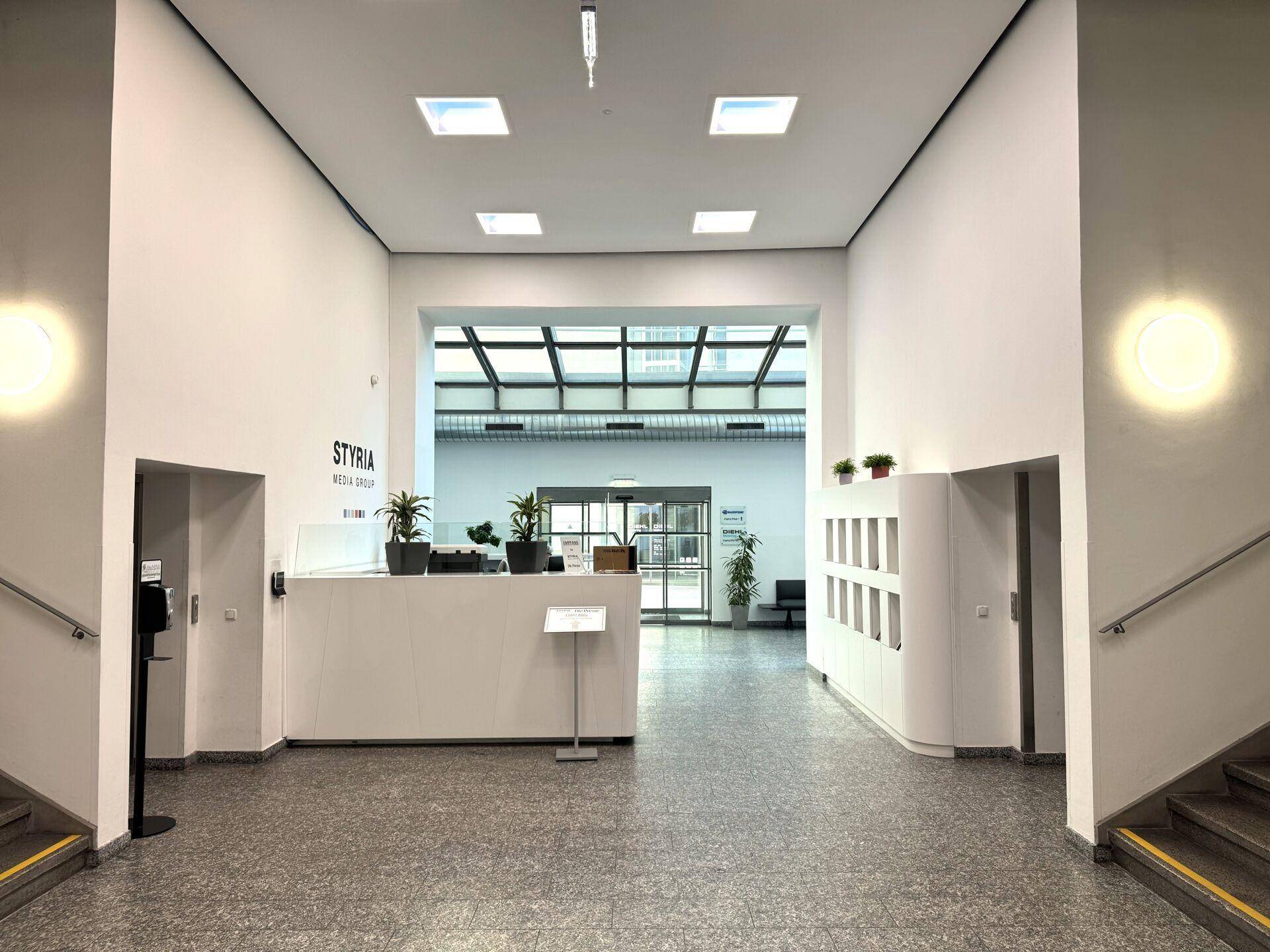 Helle und geräumige Empfangshalle mit weißem Tresen, Pflanzen und Treppen auf beiden Seiten.