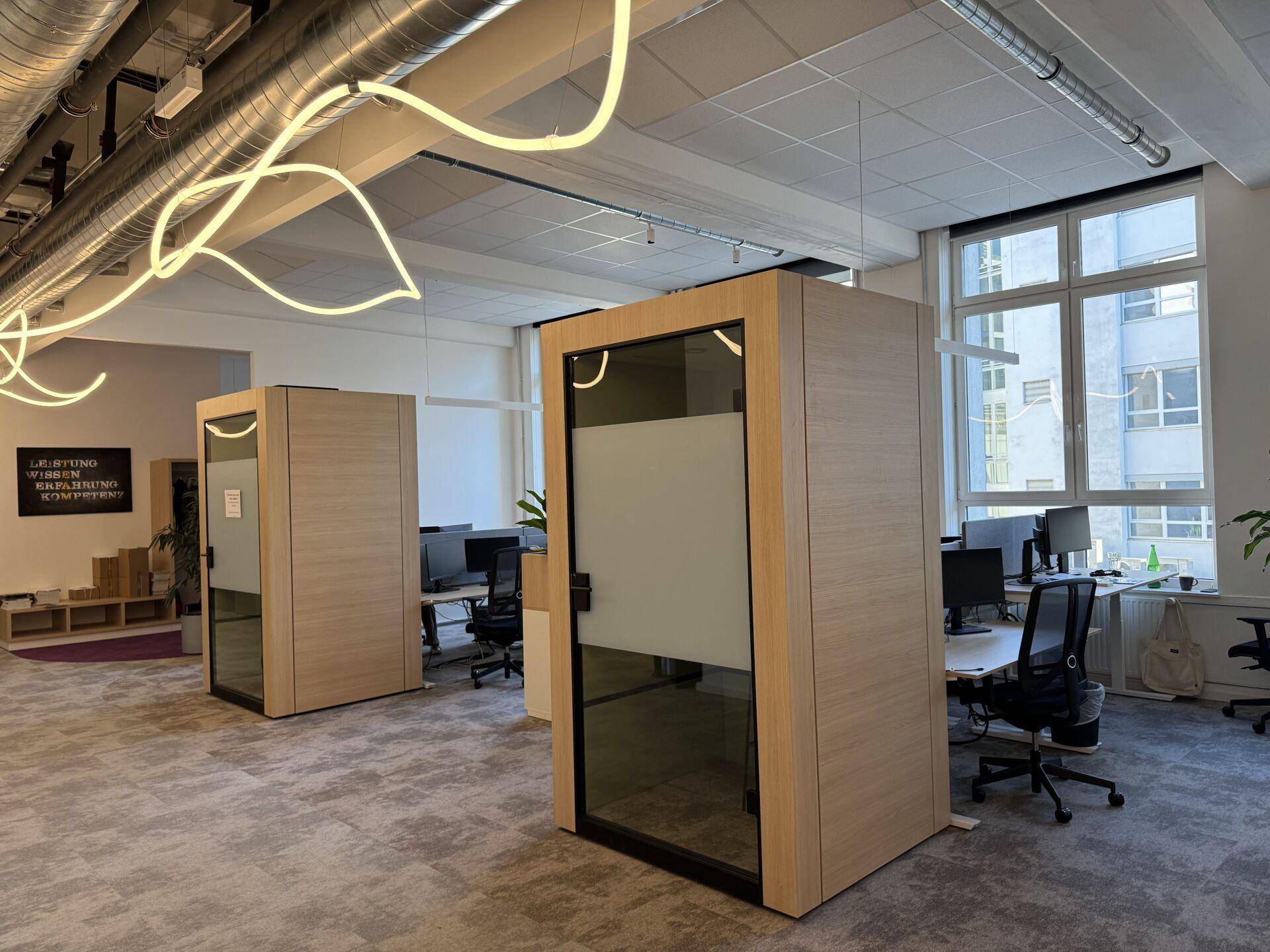 Großraumbüro mit Arbeitsplätzen, schallgedämmten Telefonzellen und moderner Deckenbeleuchtung.
