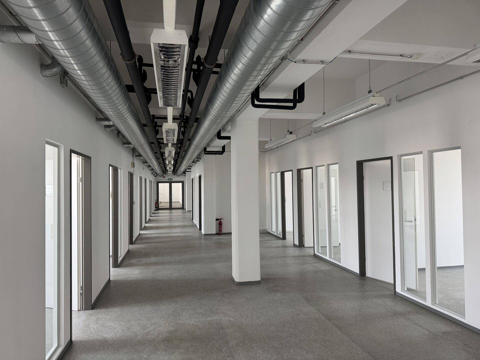 Langer, leerer Büroflur mit sichtbaren Lüftungsrohren und vielen Bürotüren mit Glasfronten.