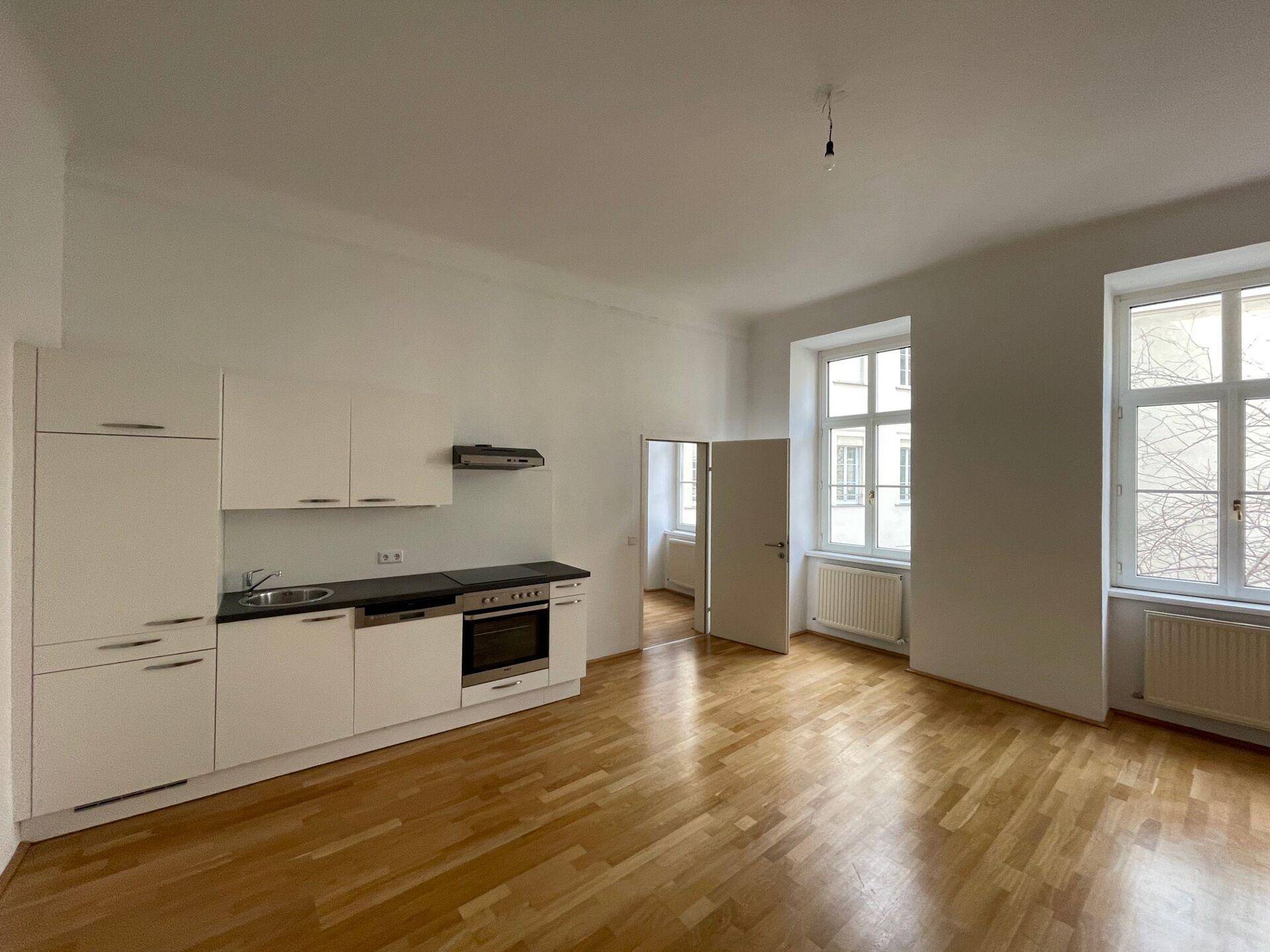 Heller Raum mit Holzboden, einer kleinen Einbauküche und großen Fenstern, die viel Tageslicht hereinlassen.