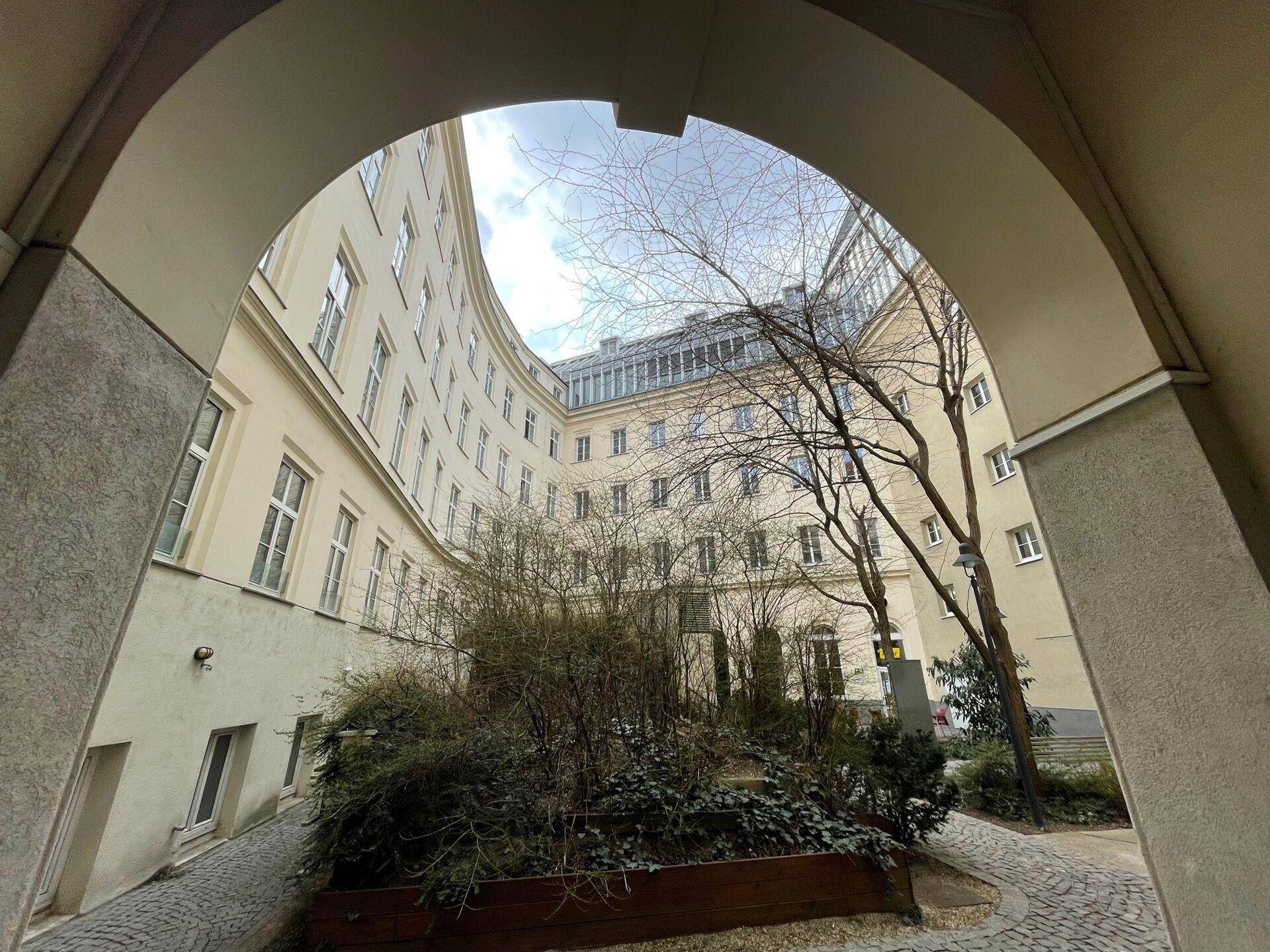 Blick durch einen Torbogen auf einen begrünten Innenhof mit Bäumen und einem geschwungenen Gebäude.