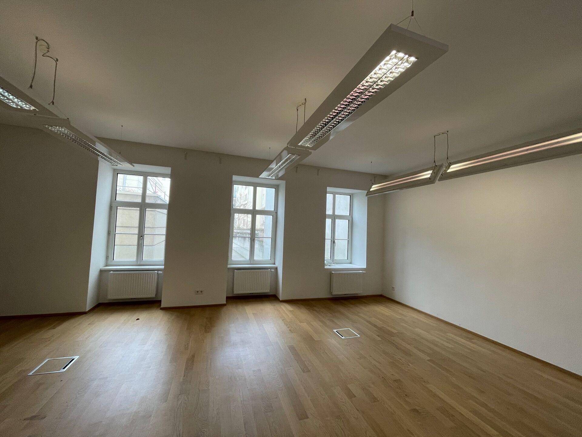 Geräumiger Büroraum mit Holzboden, großen Fenstern und modernen Deckenleuchten, bereit zur Einrichtung.