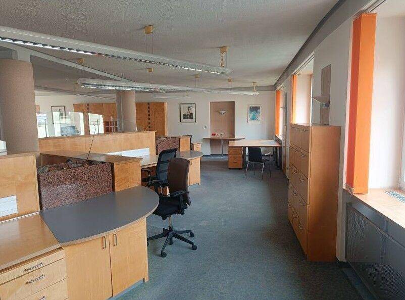 Geräumiges Büro mit mehreren Schreibtischen, Aktenschränken und viel Tageslicht durch die Fenster.
