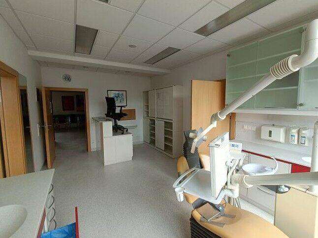 Behandlungszimmer mit zahnmedizinischer Ausstattung, Schränken und einem Empfangsbereich im Hintergrund.
