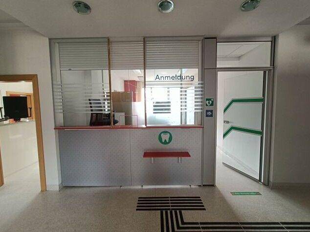 Empfangsbereich mit Schalter, "Anmeldung"-Schild und einer Tür mit grünen Akzenten.