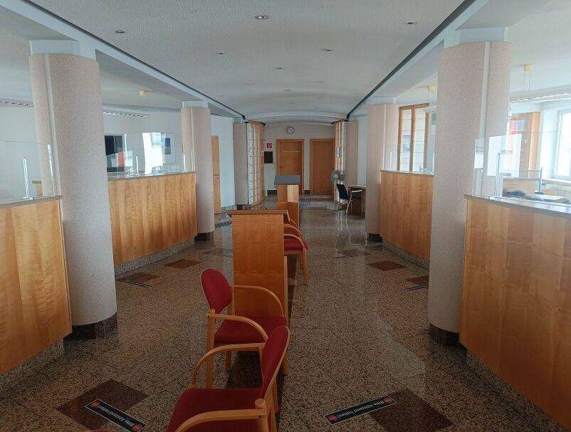 Empfangsbereich oder Wartezimmer mit Säulen, Holztresen und roten Stühlen auf Steinboden.